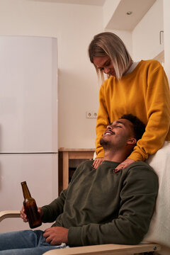 Side view girlfriend giving boyfriend massage while he is enjoying a beer sitting in armchair at home