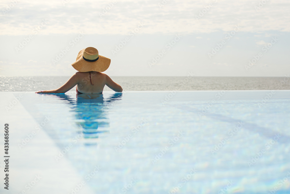 Wall mural beautiful woman in hat relaxing at edge in blue pool, enjoying summer vacation and calm sea view. sl