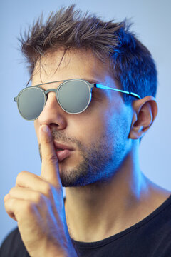 Male in trendy sunglasses showing silence sign while covering mouth with finger in studio with blue neon light