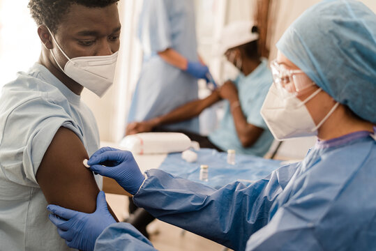Side view of female doctor in protective uniform and latex gloves disinfecting arm with cotton with alcohol to vaccinate male African American patient in clinic during coronavirus outbreak