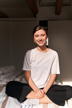 Full body of positive young lady in casual outfit and socks sitting on comfortable bed with crossed legs and smiling