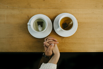 Top view of crop unrecognizable couple holding hands while sitting at table with cups of hot tea