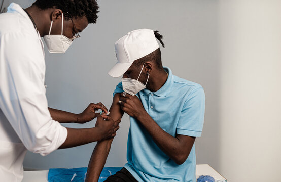 Side black doctor in protective uniform and latex gloves vaccinating male African American patient in clinic during coronavirus outbreak