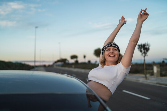 Young female in casual wear and headband with American flag print leaning out of car window and raising hands while enjoying freedom during road trip at sunset