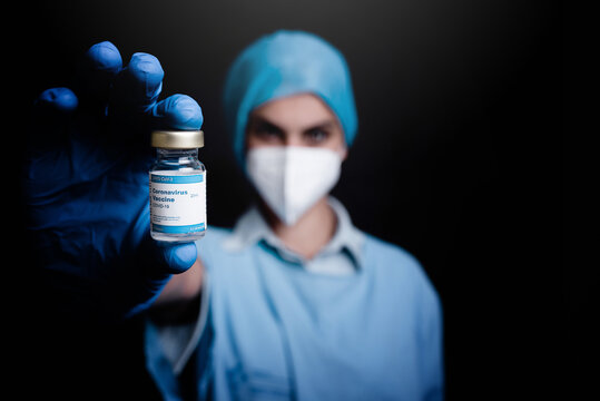 Blurred Unrecognizable Medic In Protective Mask And Latex Gloves Showing To Camera Glass Vial Of Coronavirus Vaccine While Standing In Black Background In Hospital Room