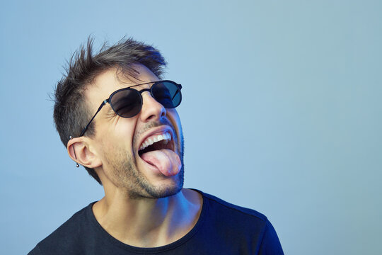 Expressive male in stylish sunglasses showing tongue and grimacing on blue background in studio with neon light