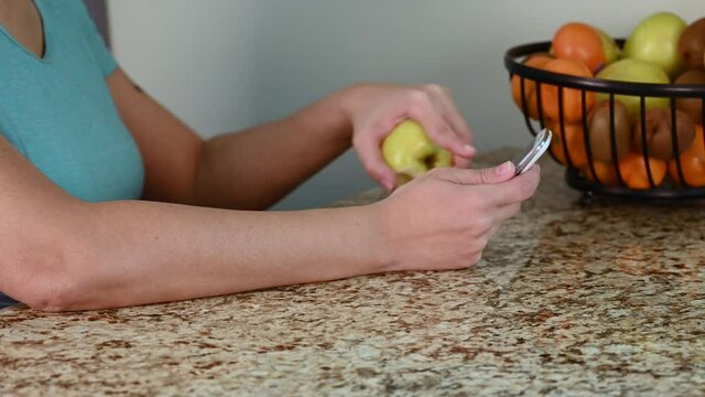 Woman checking her cell phone, texting and eating a green apple in a modern kitchen.