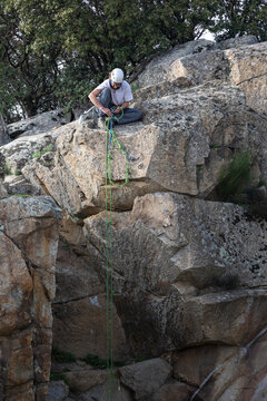 Male climber in protective helmet sitting on rock and fastening harness before descent from mountain