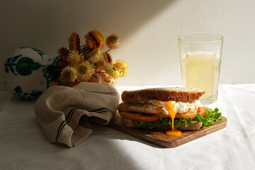 Appetizing homemade sandwich with poached eggs and fresh tomatoes and lettuce served with glass of juice for breakfast on table with flowers