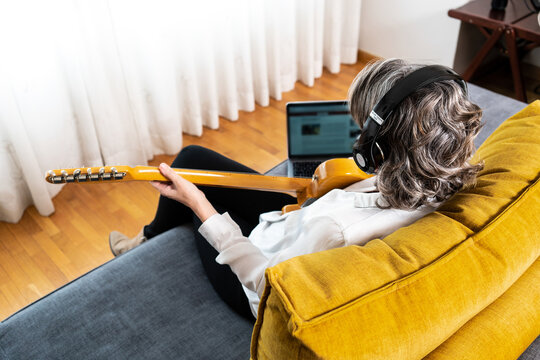 High angle back view of unrecognizable female guitarist in headphones playing musical instrument on couch with netbook at home