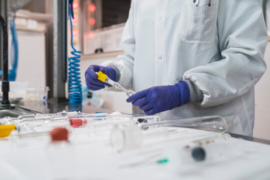 Crop anonymous competent researcher in white robe latex gloves and face mask working in modern laboratory and standing near table with assorted glassware including graduated cylinders syringes and test tubes