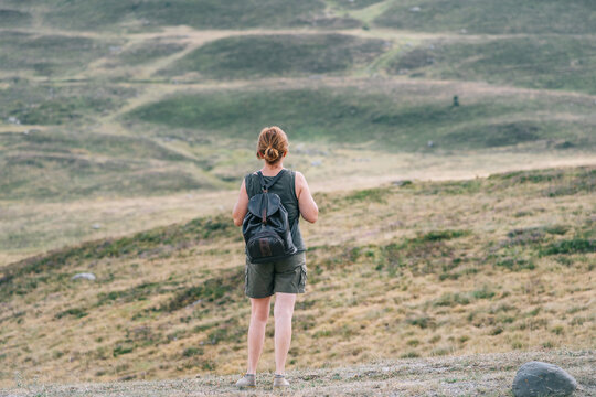 Back view of anonymous female hiker with backpack standing on hill in mountainous terrain and admiring picturesque landscape