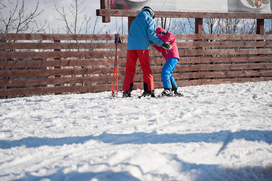 Full body faceless parent in warm sportswear and helmet teaching little kid to ski alongside snowy hill slope in winter ski resort