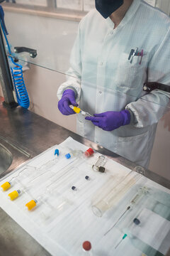 Crop anonymous competent researcher in white robe latex gloves and face mask working in modern laboratory and standing near table with assorted glassware including graduated cylinders syringes and test tubes