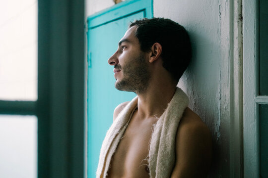 Calm shirtless male with beard leaning on shabby wall at home and looking away