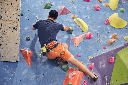 Back view of anonymous male mountaineer in harness climbing artificial wall in bouldering center