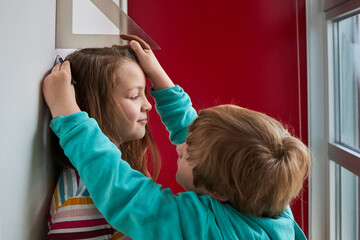 Brother helping sister with measuring her height with ruler and pencil near wall