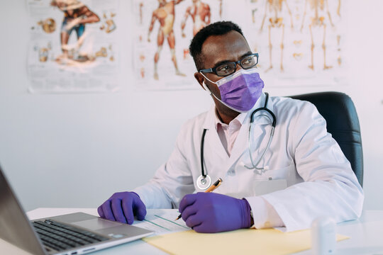 African American doctor in eyeglasses working with online patient on netbook while writing in patient file at table in hospital