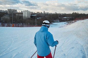 Back view anonymous male skier in warm sportswear skiing along snowy hill slope in winter city park