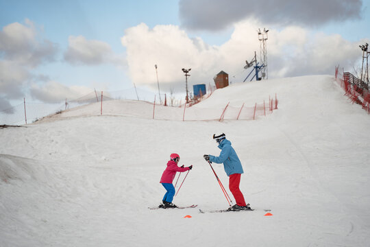 Full body faceless parent in warm sportswear and helmet teaching little kid to ski alongside snowy hill slope in winter ski resort