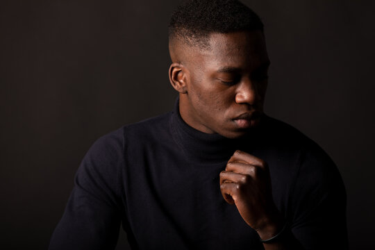 Confident young African American guy in black clothes with hand at chin closed eyes on black background in light studio