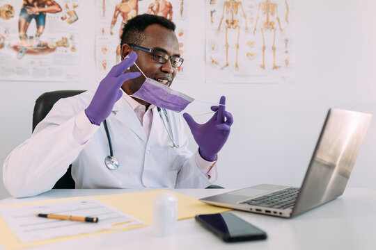 African American doctor in eyeglasses and face mask working with online patient on netbook while writing in patient file at table in hospital