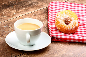 Two tasty ring doughnuts sat on a red and white checked cloth on a wooden table top with a cup of tea