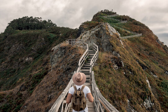 Back view of unrecognizable male tourist with backpack and hat standing on wooden pathway in a cliff and admiring scenery of mountain views in seashore during summer vacation