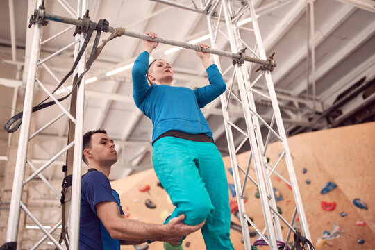 From below of female doing pull ups on horizontal bar with help of instructor while training in modern climbing center