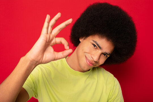 Positive African American male with Afro hairstyle smiling and looking at camera and showing okay sign while standing on red background
