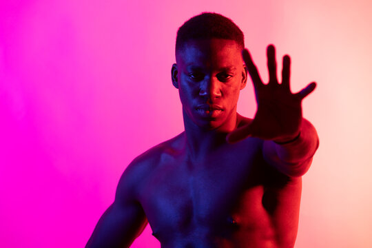 Serious young African American male athlete with naked torso looking at camera on pink background in neon studio
