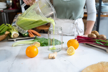 Crop anonymous female pouring tasty smoothie from blender bowl into jar at table with citrus fruits at home