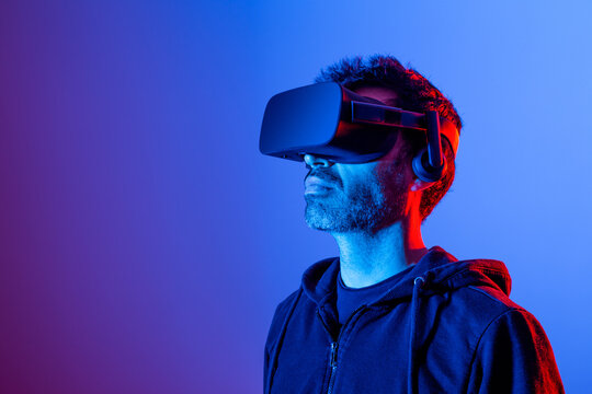 Unrecognizable bearded male in hoodie and modern headset exploring virtual reality in neon light