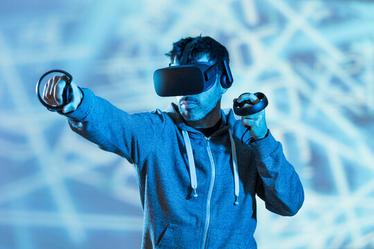 Anonymous unshaven male in hoodie and modern goggles with controllers and extended arm experiencing virtual reality