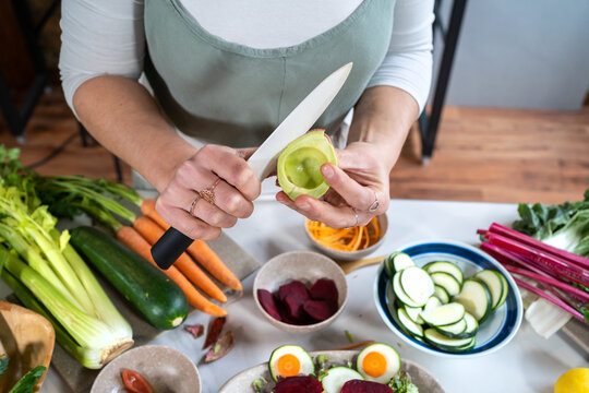 From above of crop unrecognizable person cutting ripe avocado while preparing vegetarian food with assorted vegetables