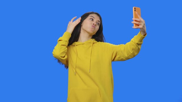 Portrait of brunette making selfie on phone, making funny faces, looking photos. Young woman in yellow sweatshirt posing in studio with blue screen background. Close up. Slow motion ready 59.94fps.