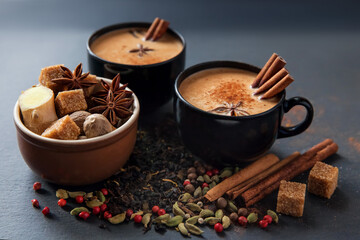 Masala tea traditional hot Indian sweet milk spiced drink in black cups. Spices cinnamon sticks, cardamom pods, star anise and coriander seeds in brown ceramic cup on a black slate stone plate
