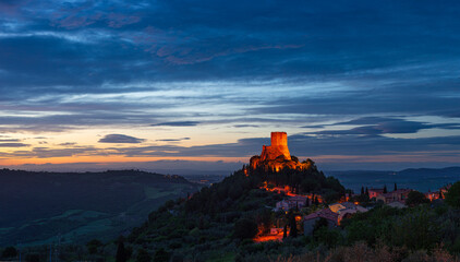 Rocca d'Oria, a medieval village and fortress in Orcia Valley, Tuscany, Italy. Unique view at dusk, the stone tower perched on rock cliff illuminated against dramatic sky.