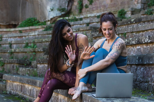 Cheerful multiracial females in activewear waving hands and having video chat while preparing for online yoga lesson via netbook