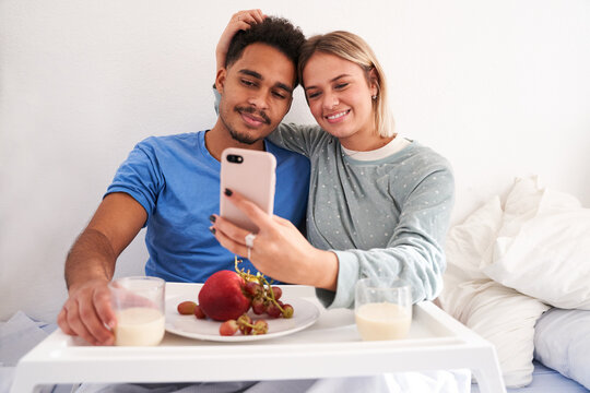 Glad multiracial couple sitting on bed and taking self shot on smartphone while having breakfast in morning - Powered by Adobe
