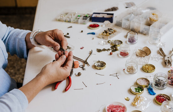 High angle of crop anonymous female master with instrument making necklace at table with different tools