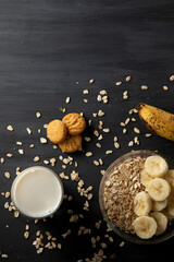 Fototapeta na wymiar Desayuno saludable de avena con plátano, un vaso de leche y unas galletas sobre la mesa de color negro.