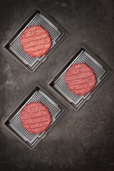 Raw Minced Steak Burgers in Shop Plastic Tray on Black Background, Overhead View. Uncooked Beef Ground Meat Patties for Grilling. Burgers for BBQ Grill in Black Container With Grill Tools, Top View.
