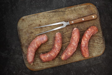 Raw Sausages In Natural Casing.  Stuffed Sausages on Old Wooden Cutting Board and Fork, Overhead View. Uncooked Sausages For Grilling or Frying on Wood Board And Black Table in Background, Top View.