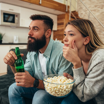 Couple drinking beer and popcorn while watching sport game at home