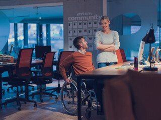 Obraz premium Businessman in a wheelchair in modern coworking office space working late night in office. Colleagues in background. Disability and handicap concept. 