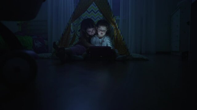 Two sisters play tablet at home. The girls are sitting in a wigwam and watching and touching the tablet screen. Video of happy children in the home room at night.