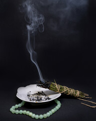 twist of dried rosemary herb burning and steaming over a white saucer on a black background. Isoteric ritual of cleansing space and spirit