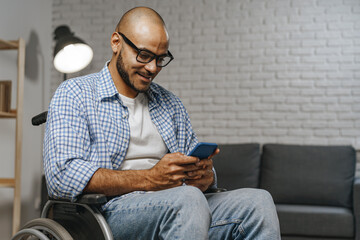 Young african american man in glasses sitting in wheelchair and using his smartphone