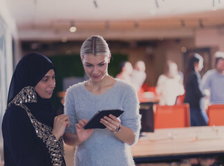 Blonde and modern Muslim businesswoman friends collaborating as a team and working on tablet. Diverse team in modern open space office in background